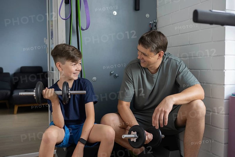 A father and son bonding during a home workout session, smiling as they lift dumbbells. The cozy setting captures their happy and active family lifestyle