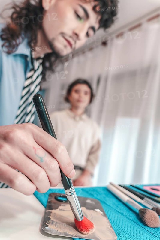Makeup artist skillfully applying foundation with a brush, carefully selecting color from a palette while a client awaits in the background, highlighting the artistry of the session