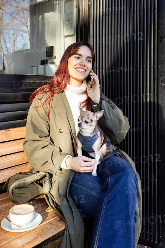 Smiling woman sits on a bench enjoying coffee with her small chihuahua in an outdoor cafe. She is on the phone, wearing a green coat and jeans, basking in the warm sunlight
