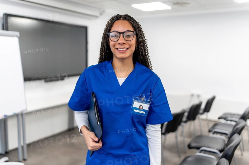 Young woman wearing a blue scrub and glasses, carrying a clipboard, smiling confidently while standing in a training room, embodying healthcare education and professional development