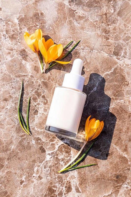 A white dropper bottle lies on a marble surface surrounded by bright yellow flowers. The minimalist design highlights a natural skincare aesthetic and beauty concept