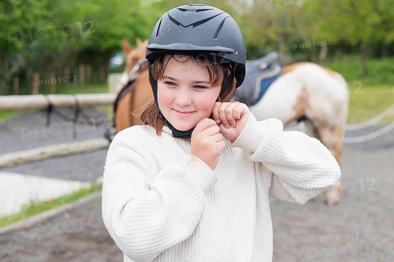 A young girl adjusts her helmet, preparing for a delightful horse ride on a sunny farm day. The serene setting and her enthusiasm perfectly capture the joy of outdoor adventures