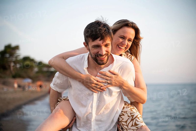 A joyful couple enjoys a playful day at an Italian beach, with one partner giving the other a piggyback ride. The serene sea and soft sand create a perfect vacation scene
