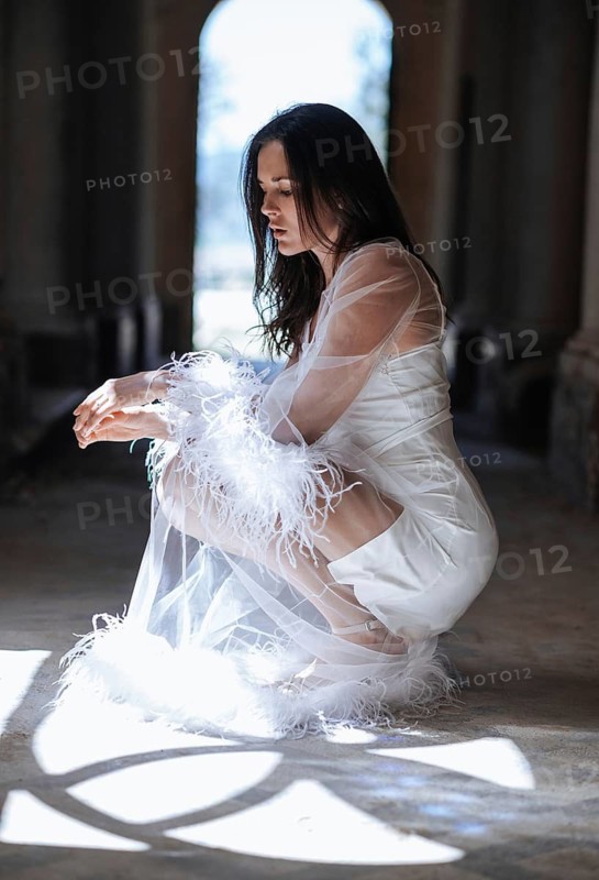 A bride in a short white gown with feather and sheer detailing kneels gracefully in a sunlit corridor of an ancient villa in Treia, Macerata. The scene evokes elegance and contemplation