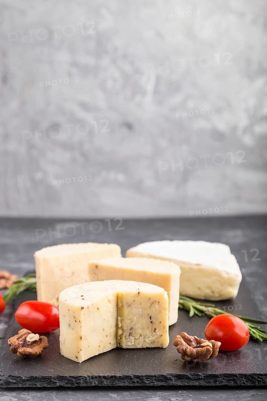 Cheddar cheese and various types of cheese with rosemary and tomatoes on black slate board on a black concrete background. Side view, copy space, selective focus