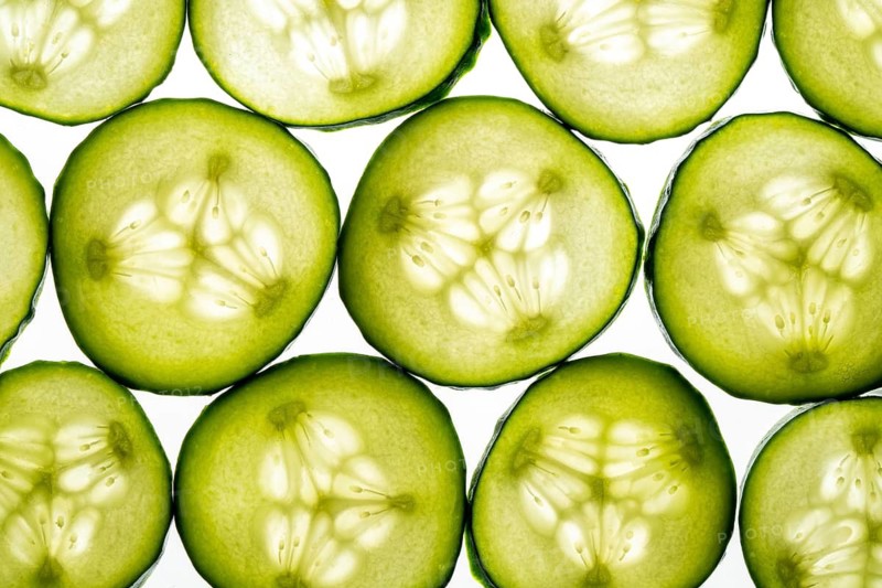 Fresh cucumber slices arranged in a seamless pattern against a crisp white background. The vibrant green hues and intricate seed patterns create a refreshing, organic design