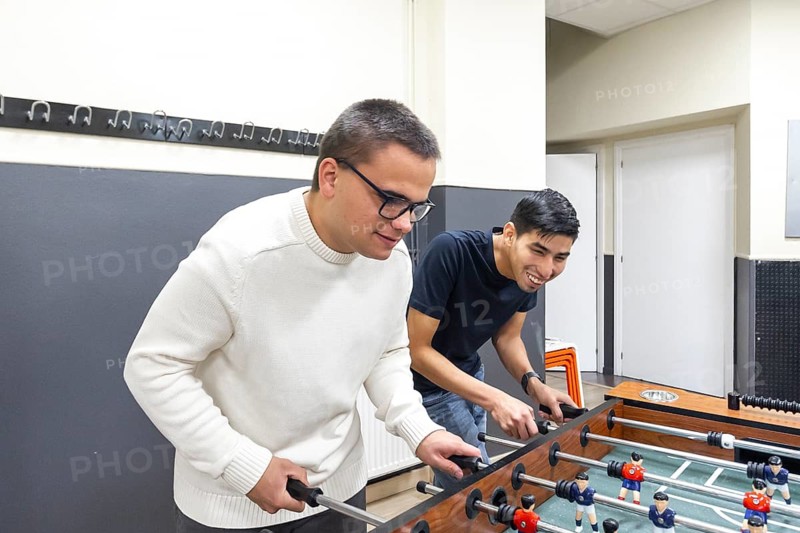 Two friends play foosball at a mental health leisure center, smiling and engaging in friendly competition. The setting promotes mental well-being through social interaction and relaxation