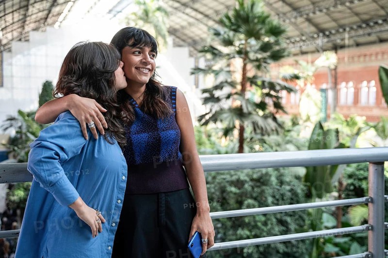 A multiethnic lesbian couple embraces at Atocha Station in Madrid. One partner lovingly kisses the other's cheek as they enjoy the lush greenery and historic architecture of the station