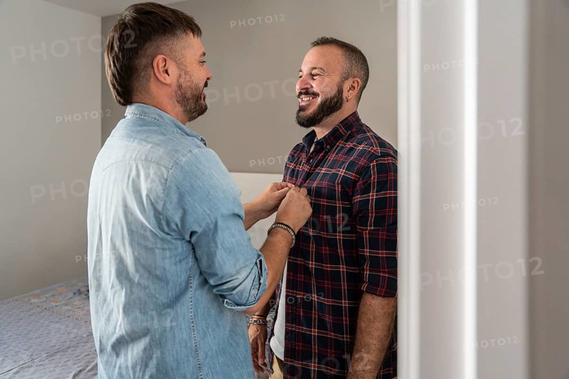 A gay couple shares a warm, joyful moment at home, smiling at each other with affection and happiness, creating an uplifting and tender atmosphere in the room