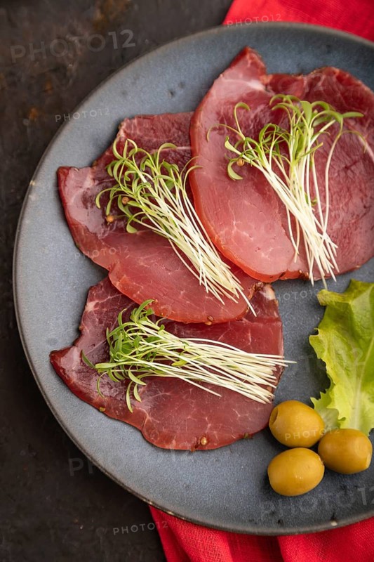Slices of smoked salted meat with cilantro microgreen on black concrete background and red textile. Top view, flat lay, close up