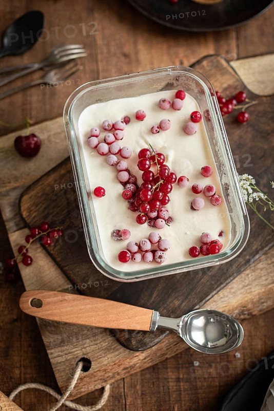 Top view of homemade vanilla ice cream garnished with frozen currants and fresh red gooseberries, served in a transparent glass dish on a rustic wooden table