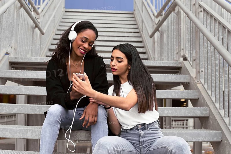 Two friends sit on outdoor stairs sharing music through headphones. One smiles while the other shows a smartphone, creating a joyful and relaxed atmosphere