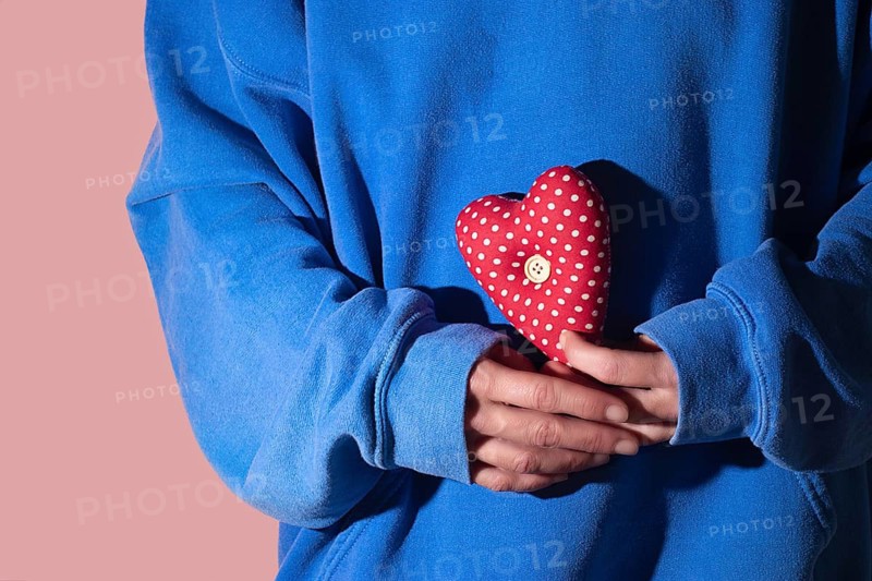 Close up of hands in blue sleeves gently holding a heart shaped cushion with red polka dots. The image conveys warmth, self love, and affection against a pink background
