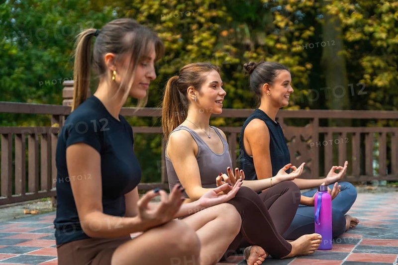 Three young women seated in lotus pose practicing outdoor meditation and mindfulness in a park, enjoying calm, inner peace, balanced breathing and healthy lifestyle together