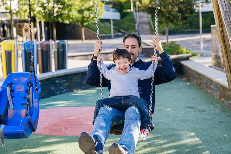 A father and his son with Down syndrome share a joyful moment on a swing in a sunny park. The child laughs gleefully, capturing the essence of pure happiness and family bonding