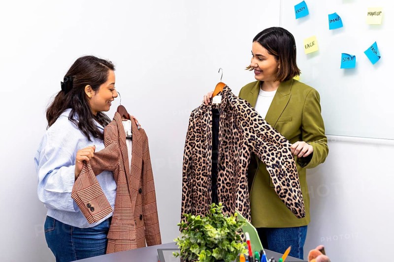 Two women engage in an image consulting session, evaluating clothing options in a modern office setting. Post it notes highlight style elements on a whiteboard, adding a creative touch
