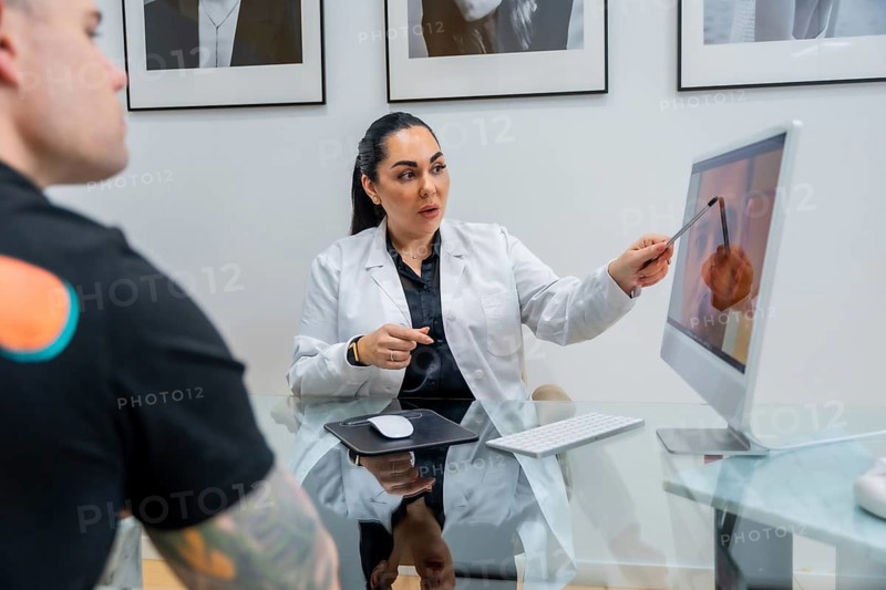 Doctor consulting male patient, pointing at computer screen with skin treatment results and cosmetic surgery options, discussing aesthetic medicine in modern clinic setting