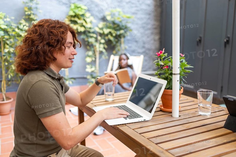 Side view of a young man works on a laptop in a vibrant outdoor coworking space, surrounded by plants and natural light, creating a peaceful and productive summer atmosphere