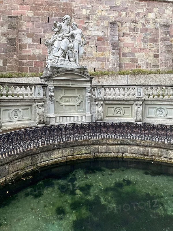 View of group of figures Mother Baar and young Danube by Adolf Heer below stone plaque with inscription Danube spring above circular spring basin designed by Adolf Weinbrenner, Donaueschingen, Baden-Württemberg, Germany