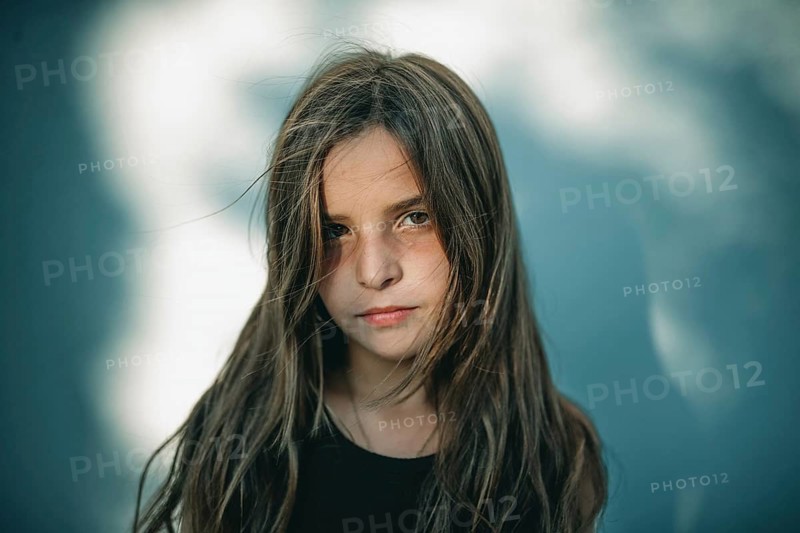A serene portrait of a teen girl standing in soft tree shadows. The gentle lighting highlights her contemplative expression, captured in natural tones