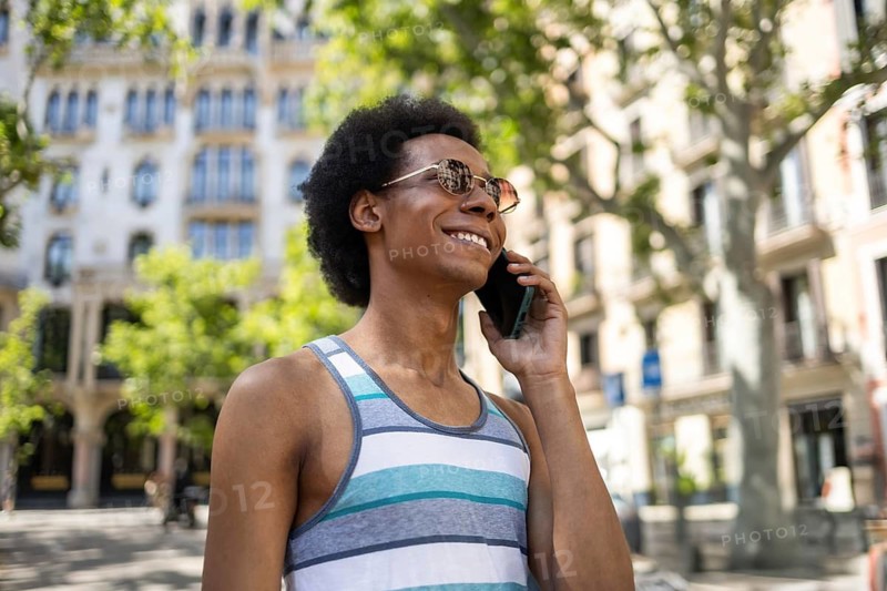 Smiling non-binary individual with afro hair enjoys a sunny day while talking on the phone, wearing a striped tank top and sunglasses, showcasing diversity and positivity