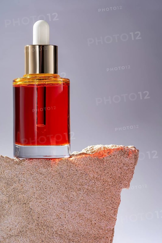 A vibrant red cosmetic serum in a clear glass bottle with a dropper, elegantly displayed on a rugged stone podium