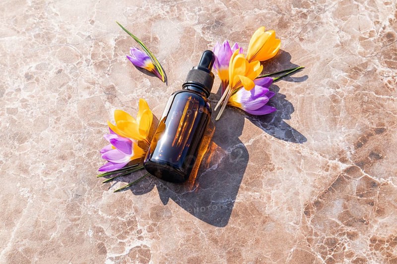 A brown essential oil bottle for skincare lies on a marble surface, surrounded by vibrant crocus flowers in shades of yellow and purple, highlighting beauty and wellness concepts