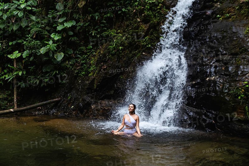 Young woman from Ecuador meditating at a serene waterfall pool in the lush Choco Andino rainforest of Ecuador, exemplifying themes of ecotourism and nature connection, capturing her peaceful interaction with the natural environment