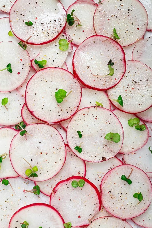 Thinly sliced radishes in vibrant red and white colors are arranged on a white surface, sprinkled with black pepper and adorned with small microgreens for a fresh look.