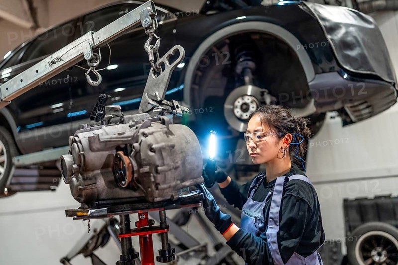 Young woman mechanic using flashlight, inspecting gearbox in car workshop with lifted vehicle
