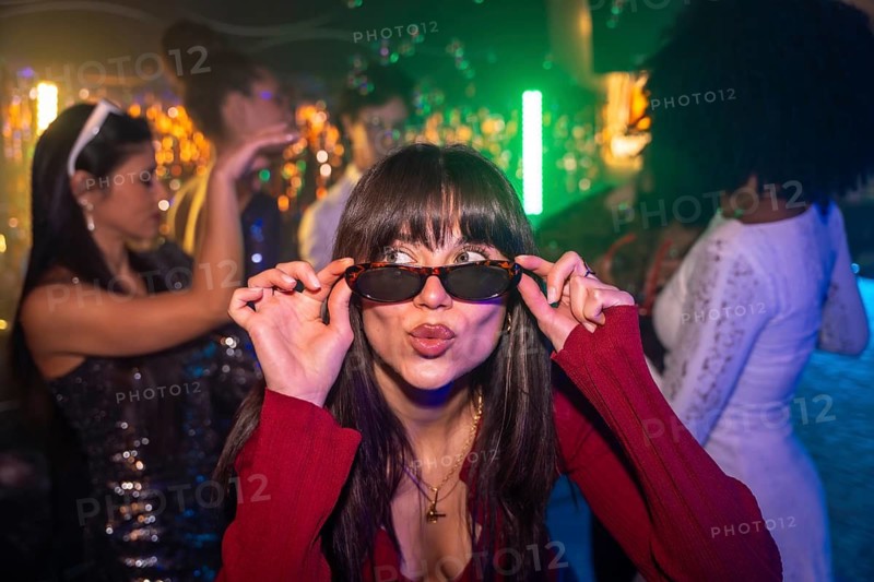 Young woman wearing sunglasses and a red outfit puckering lips, making a playful face while dancing and having fun with friends at a trendy and brightly lit nightclub party with neon lights