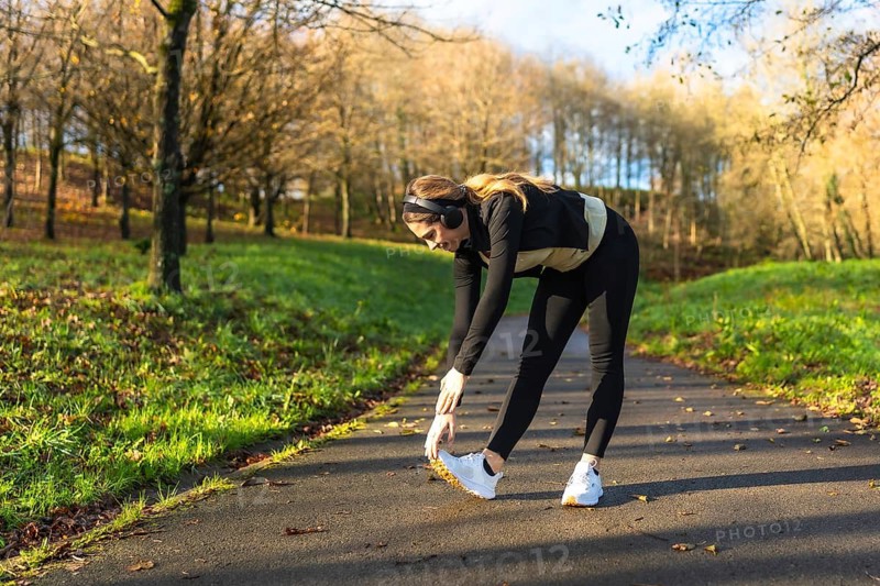Young woman with headphones performing stretching exercises on a pathway in a park, preparing her body for a healthy workout and maintaining her fitness lifestyle outdoors during autumn