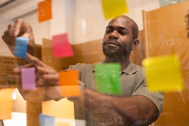 A professional in a coworking space in Barcelona places vibrant post-it notes on a glass wall. The environment fosters creativity and collaboration among diverse individuals