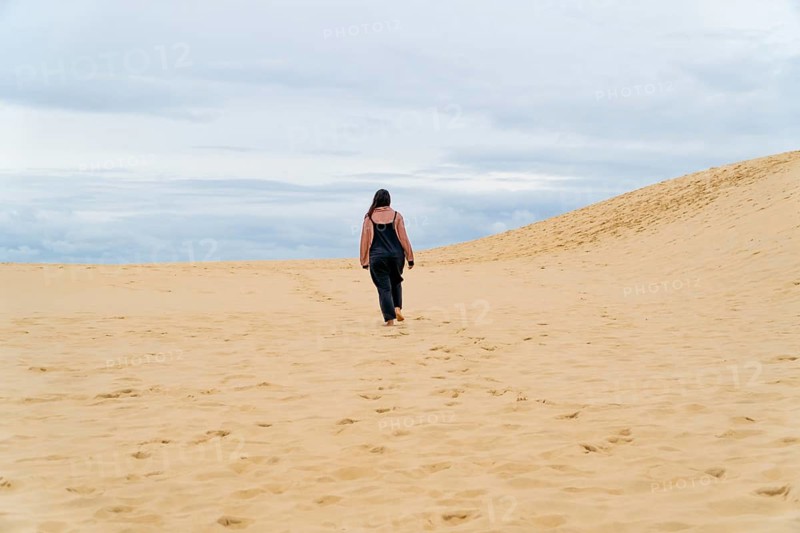 A solitary woman walks across expansive desert sand under a cloudy sky. The vast landscape and footprints create a serene and introspective scene in the dune of Pilat, France