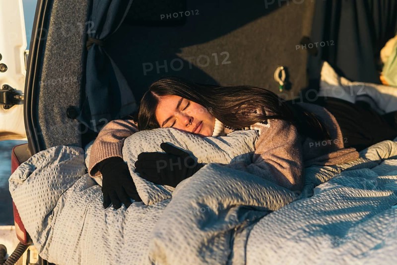 A young Latina woman sleeps soundly, wrapped in a cozy blanket in a van while camping in Iceland. The scene captures a tranquil and serene moment as the sun sets, highlighting the warmth and relaxed atmosphere of an adventurous getaway