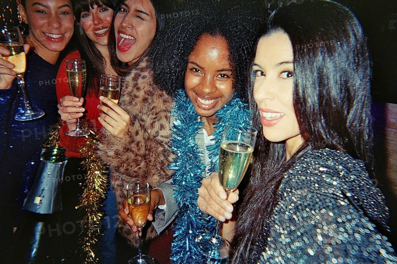 Diverse group of smiling young women friends celebrating together, holding champagne flutes and wearing festive attire with tinsel decorations during a vibrant party or gathering