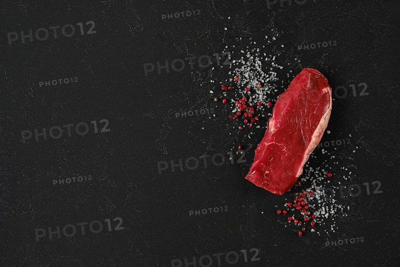 A piece of fresh raw beef sirloin steak on a dark countertop surrounded by coarse sea salt and red peppercorns, showcasing a clean and elegant presentation ideal for meal preparation