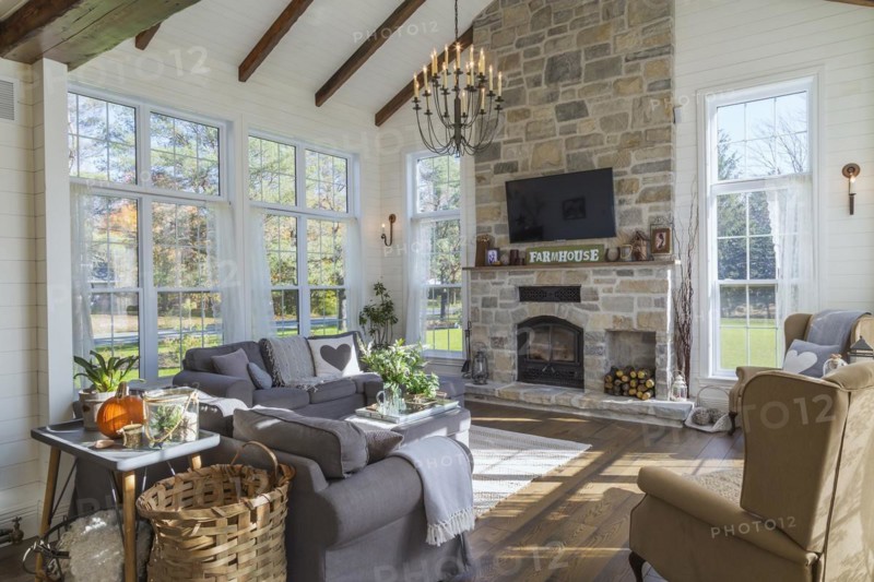 Dark grey upholstered ektorp model sofas, bergere armchairs, tan, beige and grey nuanced natural cut stone wood burning fireplace in living room with rug and stained oak wood floorboards inside country style home, Quebec, Canada