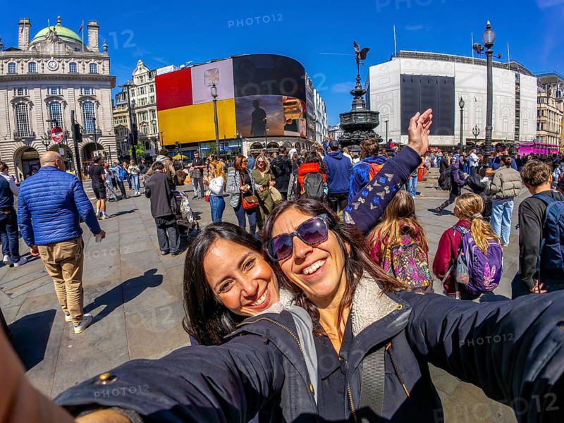 Two happy tourists taking a selfie with fisheye lens in piccadilly circus, london, with crowds of people walking in the background