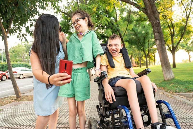 A joyful family enjoys a sunny day at the park. The girl with a tracheostomy smiles as her brother with cerebral palsy laughs in a wheelchair. They are taking a selfie together