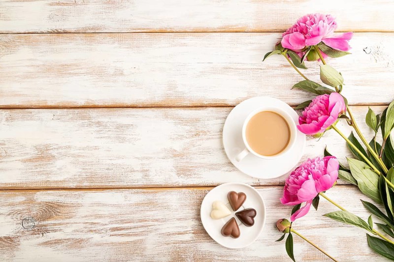 Cup of cioffee with chocolate candies, pink peony flowers on white wooden background. top view, copy space, still life. Breakfast, morning, spring concept