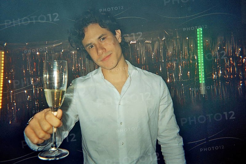 Young man smiling and holding a champagne glass, celebrating a special occasion with friends at a party, capturing candid moments with a vintage film camera aesthetic