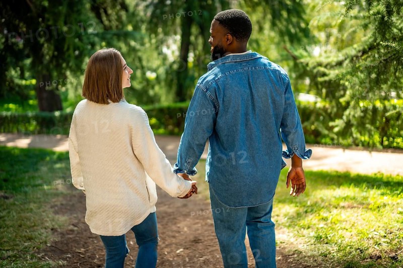 Rear view of a multi-ethnic couple walking holding hands along a park