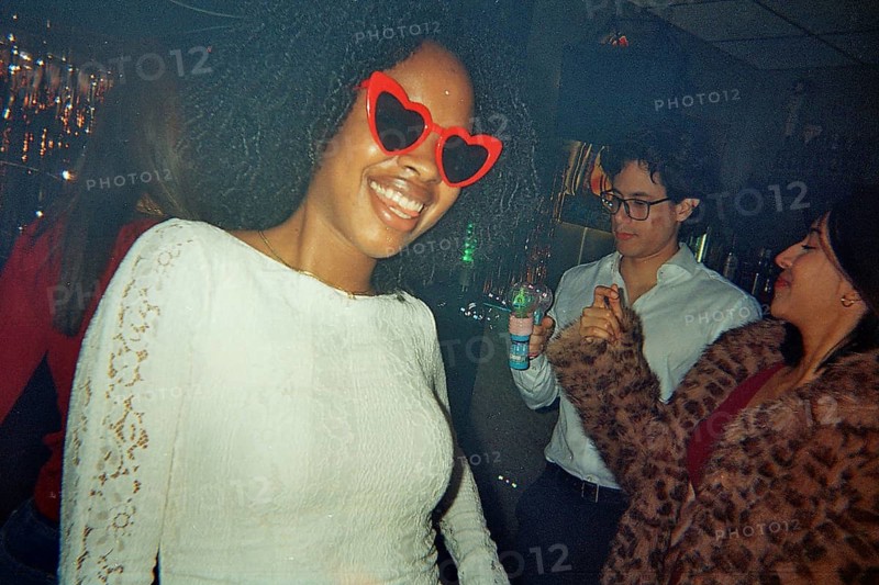 Young african american woman with big curly hair and red heart sunglasses smiling at the camera, dancing with friends at a lively party under dim lights
