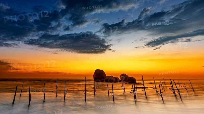 Wooden posts in the sea at sunset with dramatic clouds and orange sky, The Stilt Fishermen's Beach at Koggala in Sri Lanka