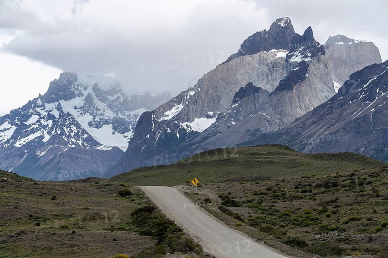 Gravel road with yellow curve signs leads through steppe landscape to the Cuernos del Paine mountain range, clouds and snowfields at the peaks, Torres del Paine National Park, Patagonia, Chile