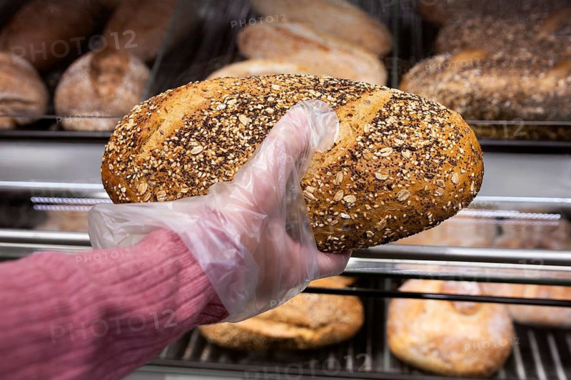 A hand in a pink sleeve uses a plastic glove to pick up a fresh multigrain loaf from a bakery display of a supermarket. The bread features seeds and oats, showcasing its homemade texture
