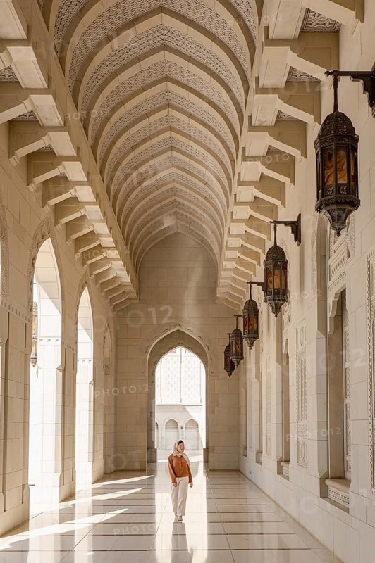 A woman in modest attire walks through the serene, sunlit corridors of the Sultan Qaboos Mosque in Muscat, Oman, surrounded by ornate arches and hanging lanterns