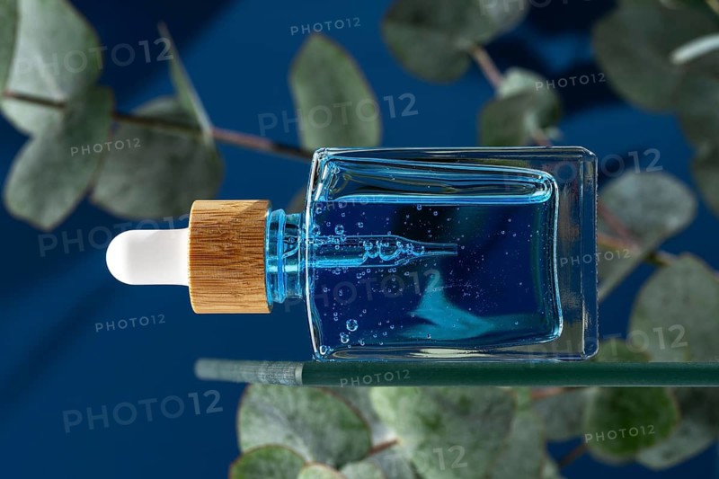 A blue face serum or oil designed for men, contained in a clear glass dropper bottle with a natural wood cap, set against a dark blue background with eucalyptus leaves