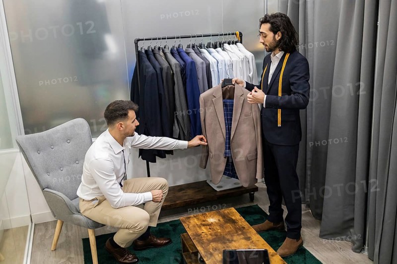 A tailor assists a customer in selecting a suit from a clothing rack in a suit store. The customer is seated, engaging attentively with various suit options